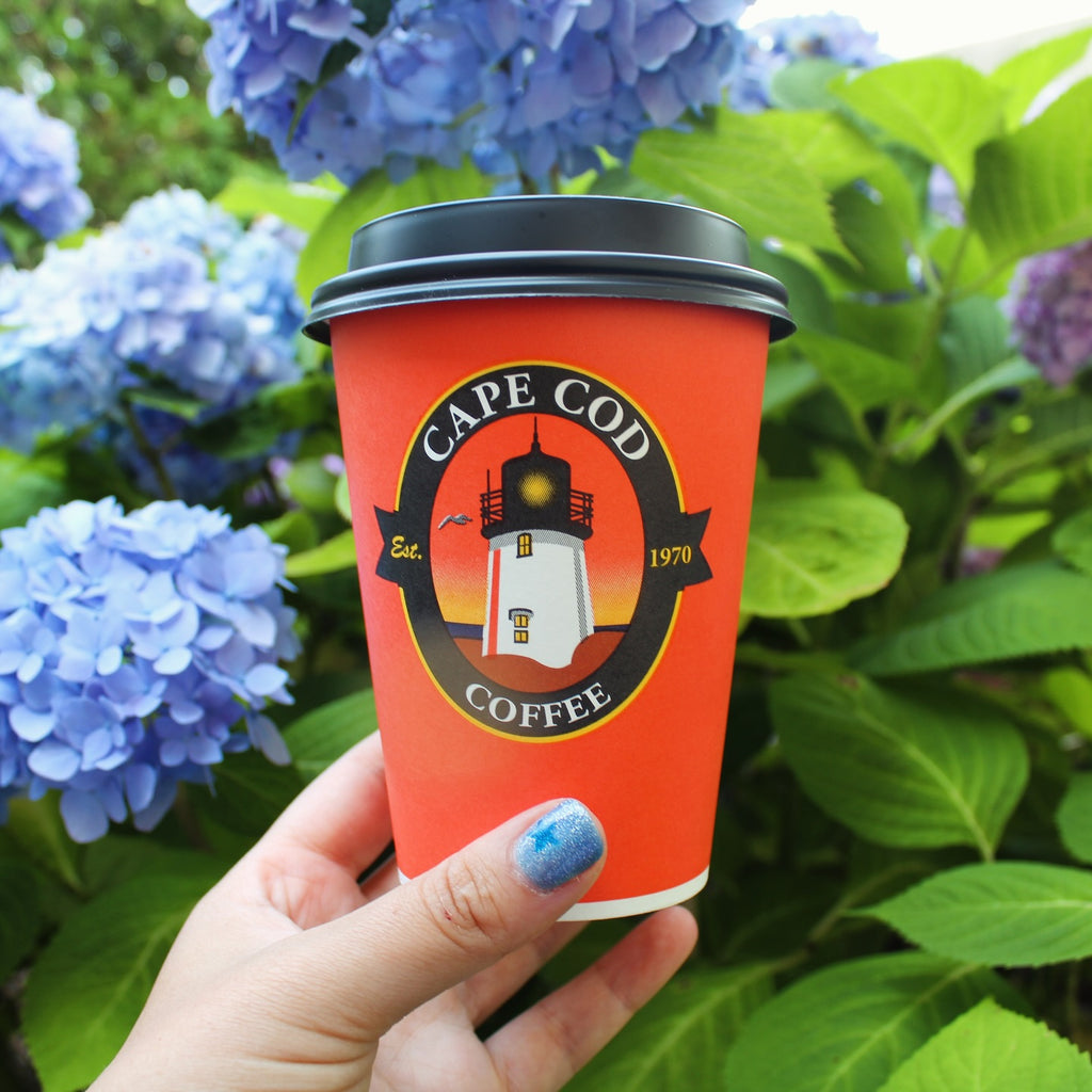 An image of an orange coffee cup being held in front of blue hydrangea flowers. The cup is orange with a black lid and a colorful lighthouse logo that reads "Cape Cod Coffee, Est. 1970"