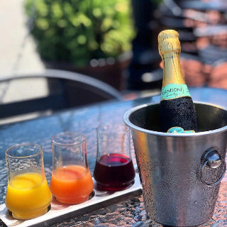 Bottle of Candoni sparkling wine in a metal ice bucket with three glasses of juice on a table outdoors.