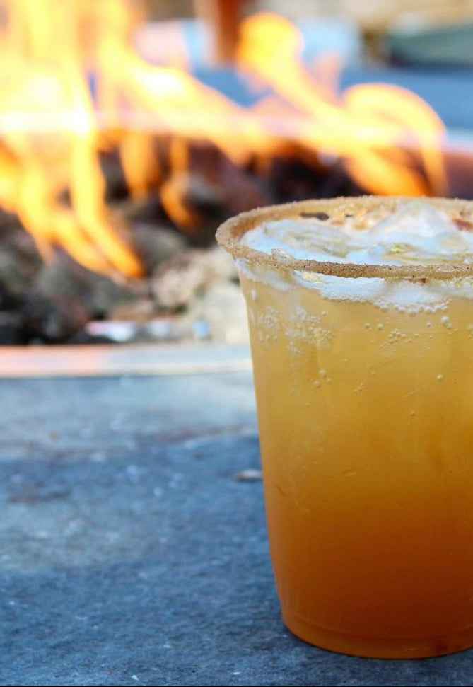 An image of an orange cocktail in front of a fire pit.
