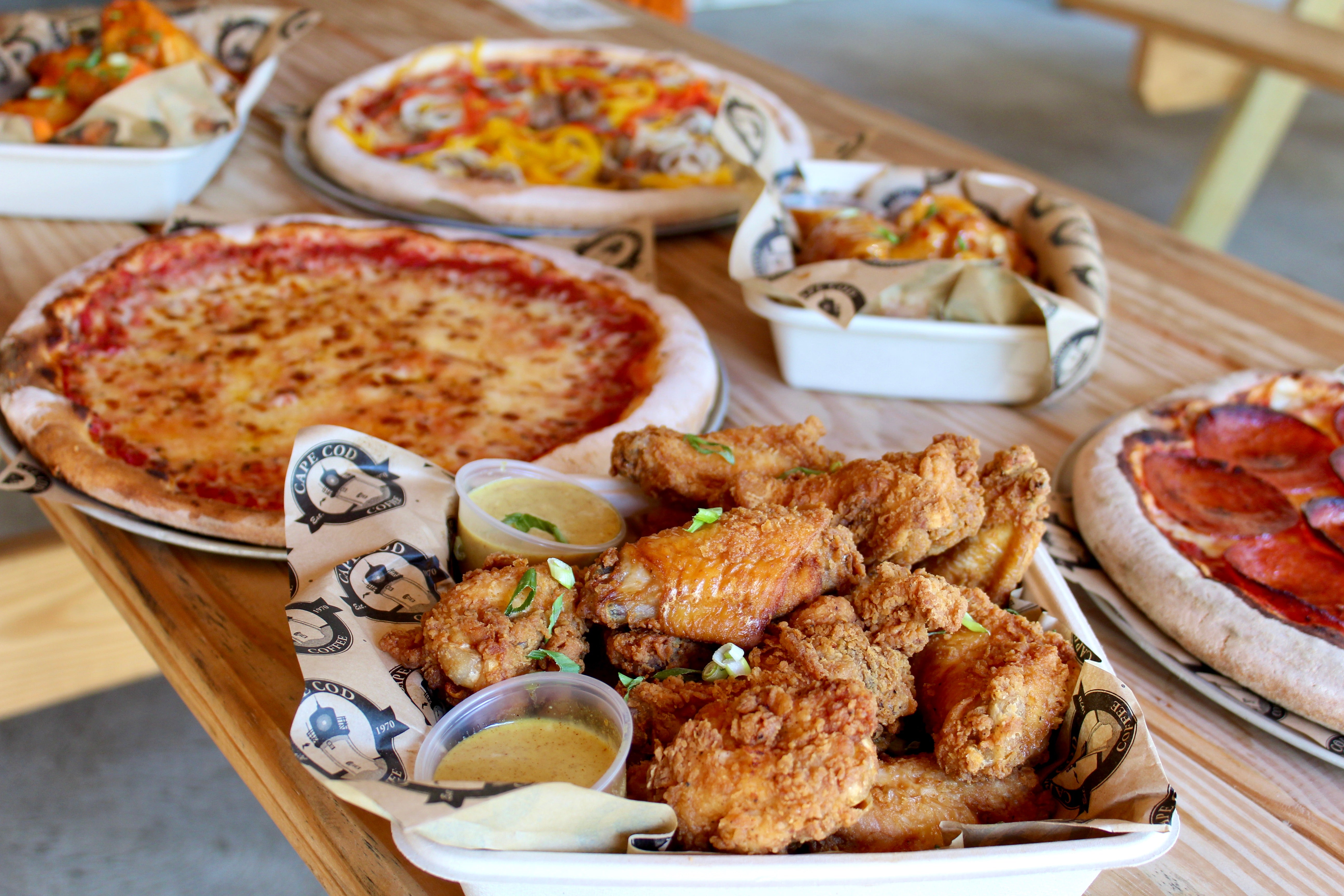 A photo of several food dishes ona wooden bench. The closest to the camera is a basket of chicken wings with honey mustard sauce. Behind it is a pepperoni pizza, more wings, a cheese pizza, a pizza with peppers, and buffalo wings.