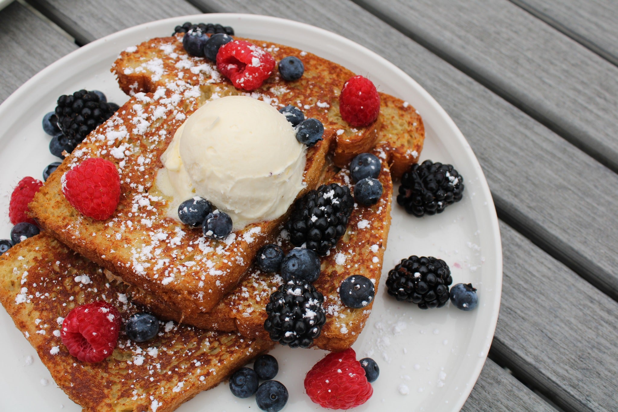 An image of french toast on a white plate that has butter, raspberries, and blueberries on it.