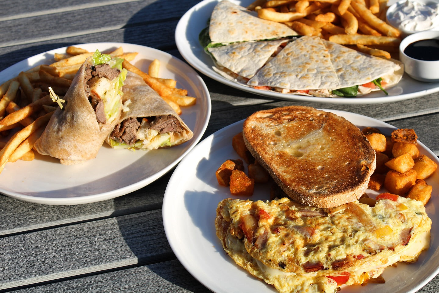 An image of three dishes on a wooden table. The closest to the camera is an omlette with peppers, cheese, and bacon in it, served with toast and hasbrowns. The second is a wrap with lettuce, meat, and cheese served with french fries. The third, farthest away, is a quesadilla with french fries.