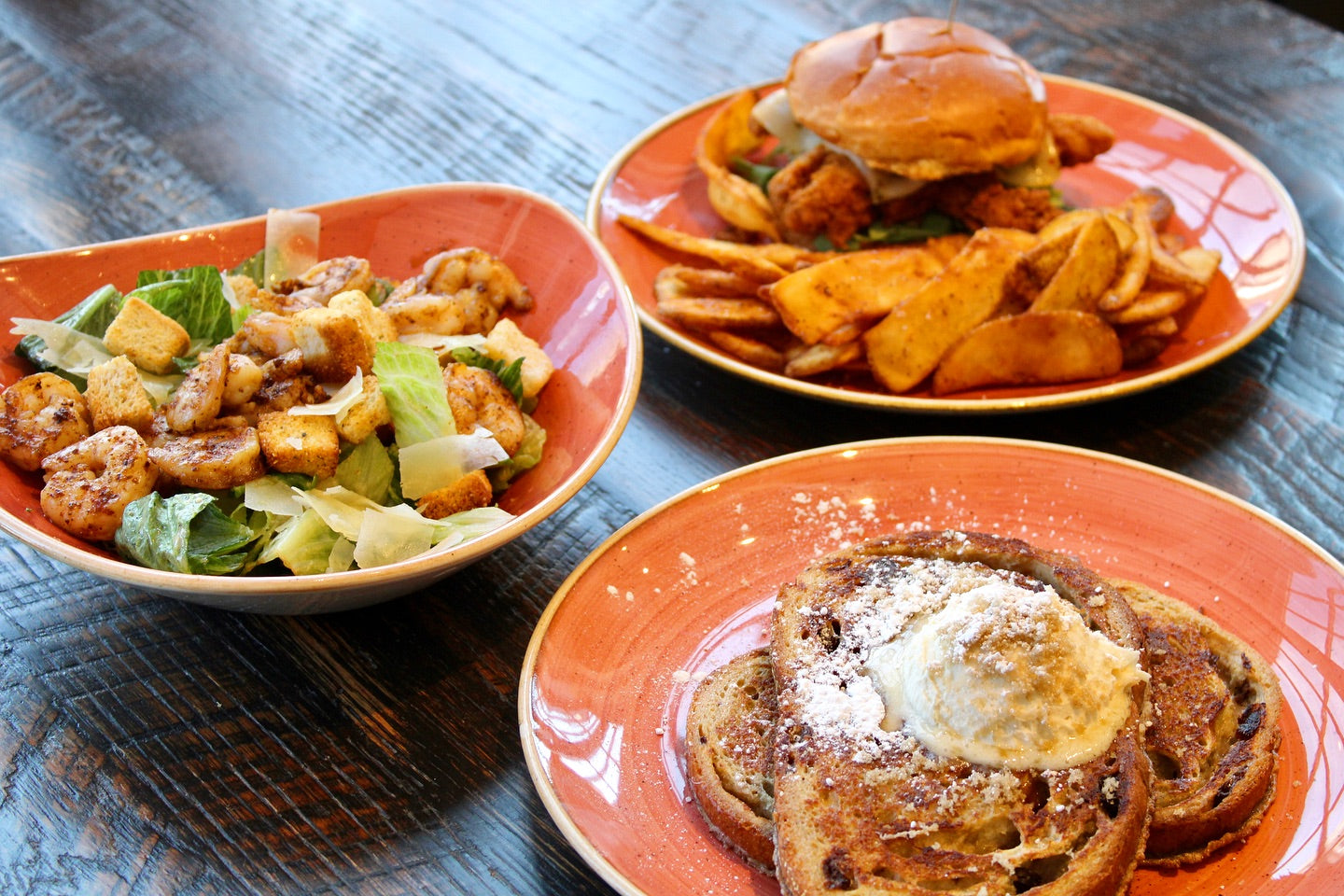An image of three dishes on a wooden table. The closest to the camera is a plate of french toast with butter and powdered sugar. The second is a Caesar salad with croutons. The farthest away is a crispy chicken sandwich with french fries.