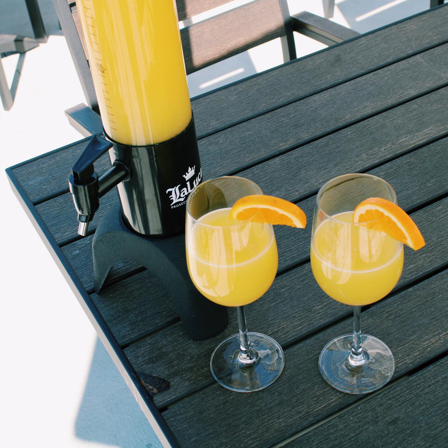 A black wooden table that has a tall container of mimosa in it. Next to it are two wine glasses filled with mimosa with two orange slices on them.